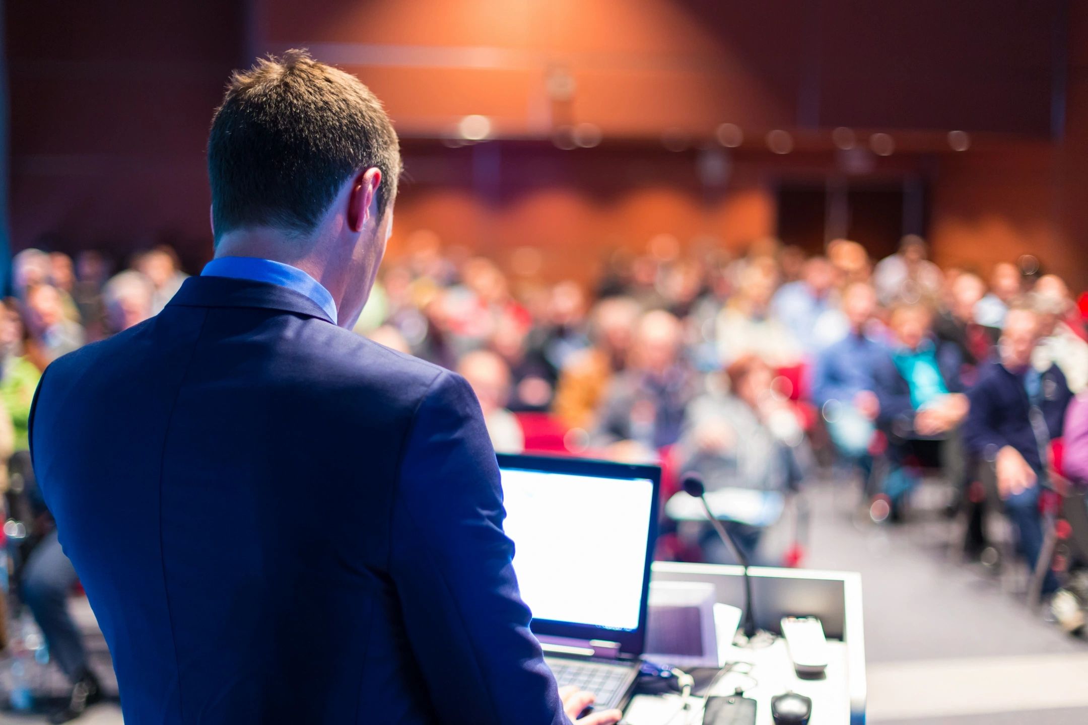 man looking at laptop with a lecture theatre full of people in the backgrouind